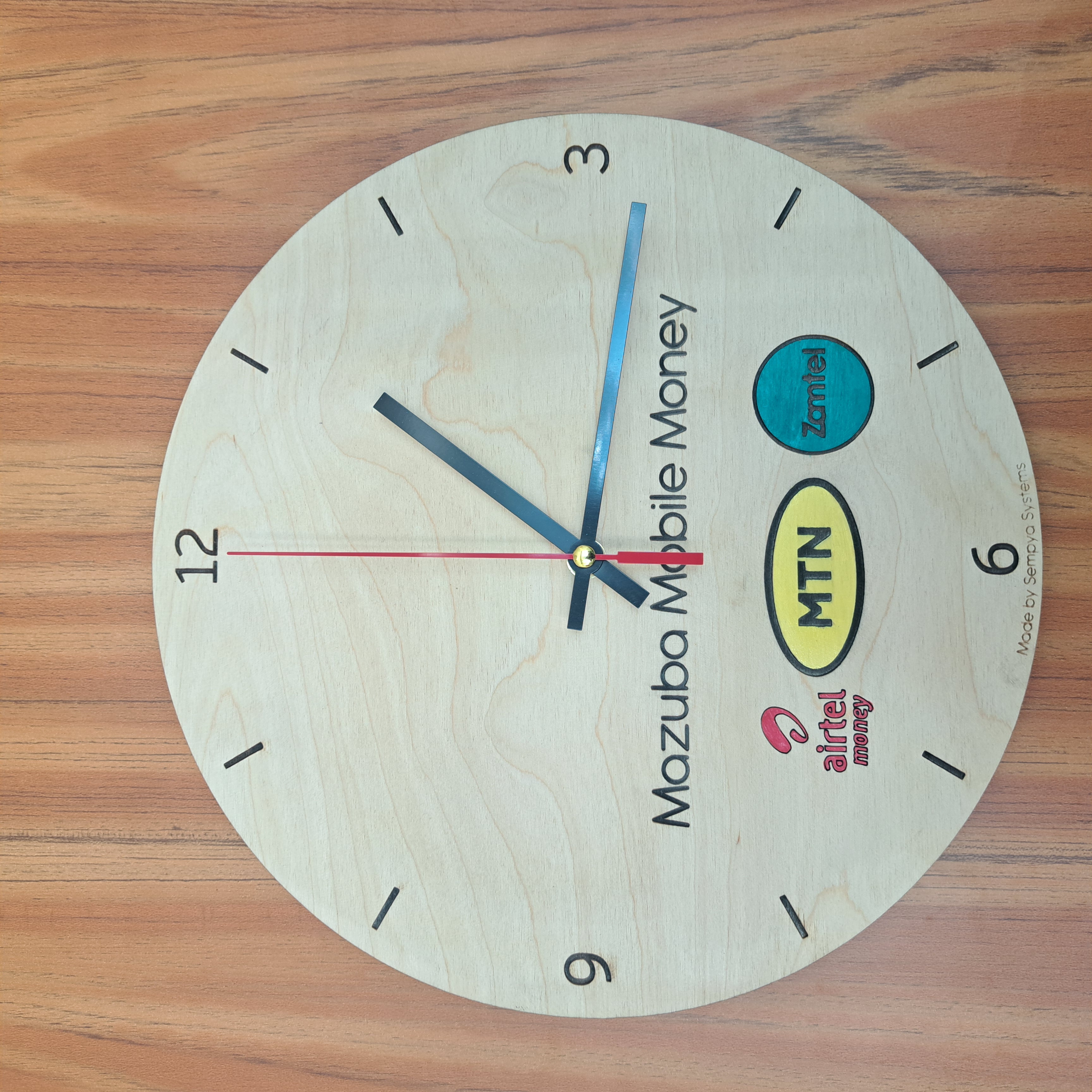 Laser-engraved plywood wall clock for Mazuba Mobile Money with hand-painted Airtel, MTN and Zamtel logos